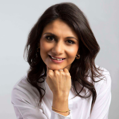 A professional headshot of Anisha Patel facing the camera directly against a plain white background. They have long dark brunette hair falling naturally over their shoulders, smiling with an open-mouthed smile and their chin is resting on their fist. They are wearing a white shirt, a thin gold bangle, a gold circular fan-shaped earring visible on the right side, and a ring on their ring finger.