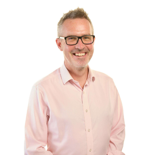 A professional headshot of Kevin Monahan with their shoulders angled slightly to the right and their face turned toward the camera. They have short blonde hair, black-framed glasses, light beard stubble, and the photo captures a broad smile and a joyful expression. They are wearing a light pink shirt with the top button undone.