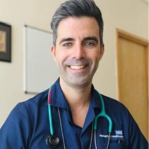 A headshot of Filipe Carvalho facing the camera directly, shown from the shoulders up. They are smiling broadly with their upper teeth visible and have grey hair styled in a quiff. They are wearing a dark blue NHS healthcare uniform with an NHS logo visible on the chest pocket, a pen in the pocket, and a stethoscope around their neck. The background includes a black frame on the far left, a whiteboard in the centre, and an oak door on the far right.