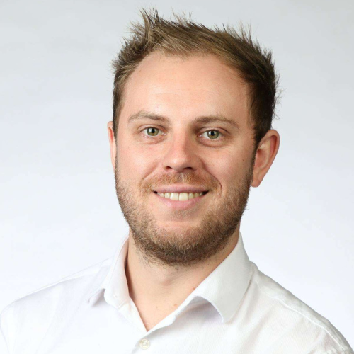 A professional headshot of Joseph Williams with their shoulders angled slightly to the left and their face turned toward the camera against a plain white background. They have short, straight blonde hair, light blonde beard stubble, and a slight smile. They are wearing a plain white shirt with the top button undone.