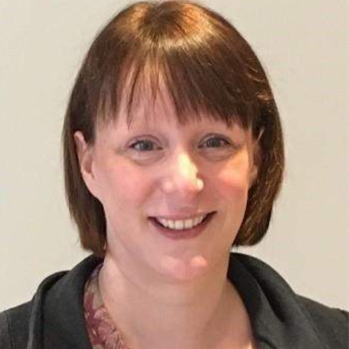 A headshot of Eva Morris facing the camera directly, shown from the collarbone up. They are smiling warmly with an open-mouthed smile and have dark brunette hair styled in a bob with a fringe. They are wearing a black collared jacket over a maroon top featuring a leaf pattern in green and light pink.