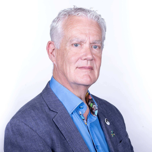 A professional headshot of Mark Saunders against a plain white background, with their shoulders angled slightly to the right and their face turned toward the camera. They have short white hair and a neutral expression with a slight smile. They are wearing a navy blazer with two badges on the left lapel, including the Star of Hope badge for Bowel Cancer UK. Under the blazer, they are wearing a light blue shirt with the top button undone, revealing a colourful abstract pattern on the inside of the collar.