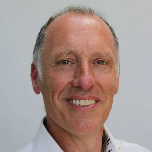 A professional headshot of Richard Adams, shown from the shoulders up against a white background. They have short grey hair and are smiling broadly, with their upper teeth visible. They are wearing a white shirt featuring a green patterned lining on the inside of the collar.