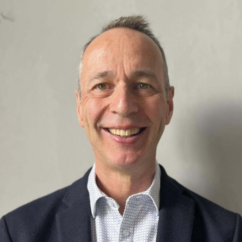 A headshot of Richard Wilson facing the camera directly against a plain white background. They have short grey hair, a broad smile with their upper teeth visible and a joyful expression. They are wearing a navy blazer over a blue-and-white checked shirt, with the top button undone.