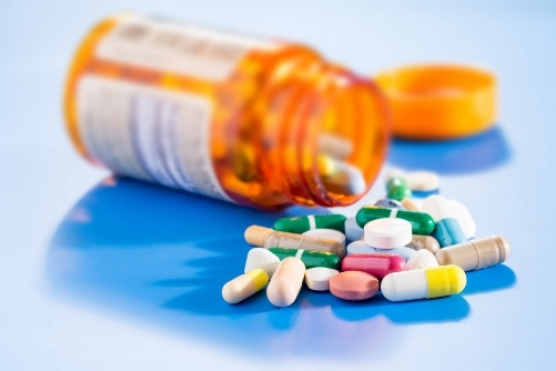 A photo of an orange pill bottle on it's side with a white label on it. From the bottle are spilling a selection of pills in various colours, with some still in the bottle. The bottle and pills are on a light blue table and you can see the orange pill bottle lid, out of focus, in the background.