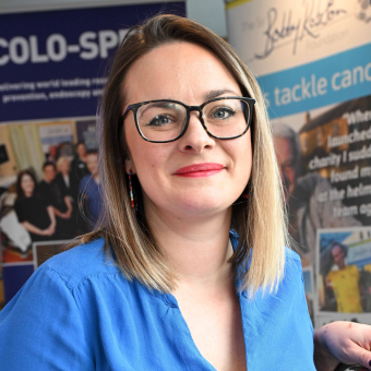 A photo of Dr Christina Dobson smiling at the camera. She is wearing black framed glasses and a blue blouse. Behind her are two pull up event banners.