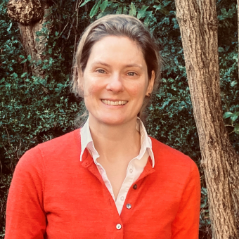 A photo of Professor Katie Robb. She is stood in front of some trees and other greener and is smiling at the camera. She has her hair tied up and is wearing a red jumper with a white collared shirt underneath.
