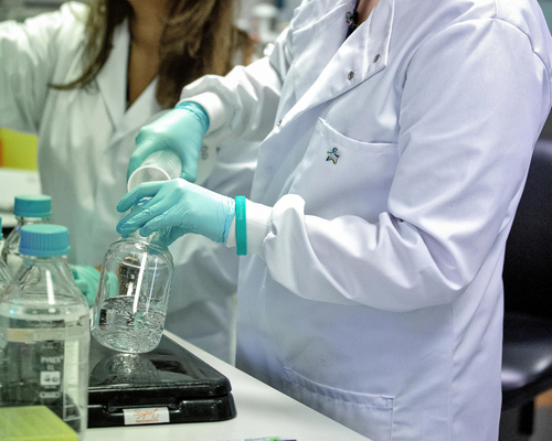 Two scientists work in a laboratory wearing white lab coats. In the foreground, the scientist closest to the camera wears a Star of Hope badge and a light teal Bowel Cancer UK wristband. Using gloved hands, she pours a clear liquid from a measuring cylinder into a glass beaker on a black scale. Several bottles with light blue lids containing clear liquid sit on the workbench. A second scientist stands beside her in the background.