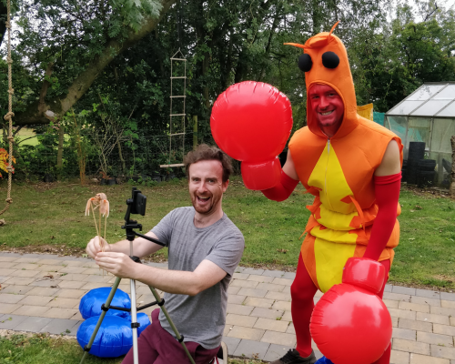 A photo of Shane and Liam in a garden. Shane is on the left of the image and is kneeling down behind a tripod holding a phone. He is holding a crustacean which is attached to some cocktail sticks. He is wearing a grey t-shirt and red trousers. Liam is on the right of the image and is dressed up as a crustacean. His face is painted red and he has big inflatable red boxing gloves on both of his hands. He is waving to the camera with his right hand. They are both smiling at the camera.