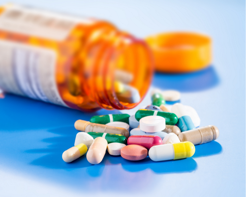 An image of an orange pill bottle open on it's side on a blue table with a selection of multi-coloured pills coming out. The orange lid is visible and out of focus in the background