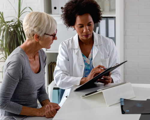 A healthcare professional is sitting opposite the camera in a white coat holding a black clipboard. To the left of the image a patient is sat in a grey top listening to the healthcare pofessional. Documents and a tablet can be seen on the white table they are sat at, in the background a large indoor plant is visible.