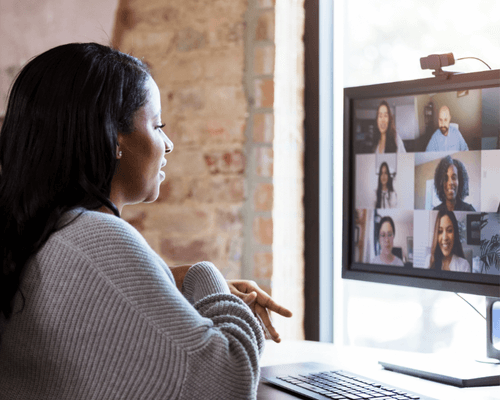 An image of a woman sitting at a desk in profile with an exposed brick wall behind them. The desk has a computer on it with a video conference call going on