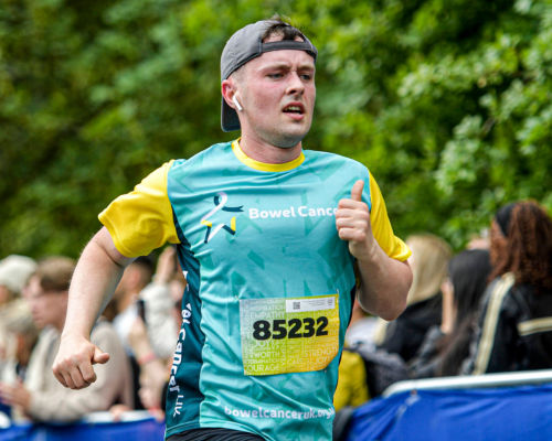 A photo of a runner in a Bowel Cancer UK branded technical t-shirt running past cheering spectators, behind them out of focus spectators and green trees can be seen.
