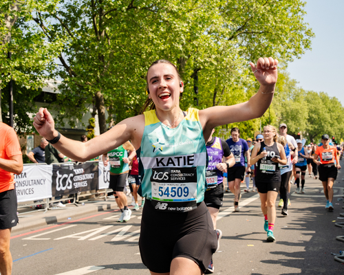 A photo of a woman running the London Marathon wearing a Bowel Cancer UK branded running vest. They are smiling into camera and have their arms out to either side. Other runners and trees are visible down the road behind the runner, it is a sunny day.