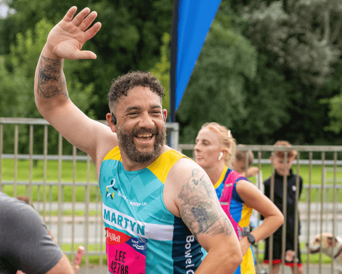 A photo of a runner smiling and waving at the camera. He is wearing a light teal branded running vest. In the background there is a blue flag banner, a metal fence and other runners.