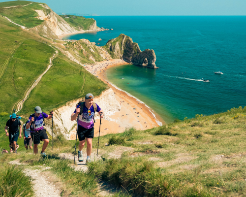 A photo of people taking part in the Jurassic Coast Ultra Challenge. The image shows a small group of people climbing up a small hill. The background shows a view of the sea.