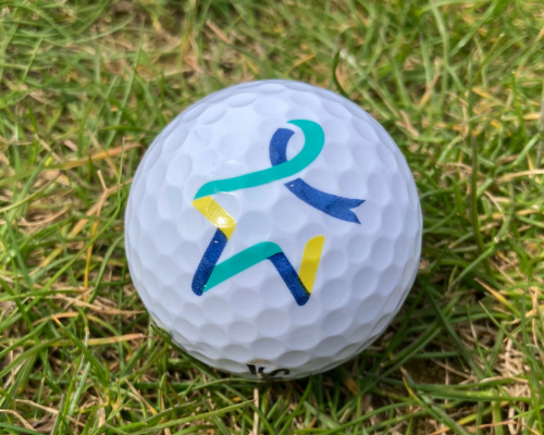 A close up photo of a golf ball. The golf ball is sitting on some grass. It is white, with a light teal, dark teal and yellow Star of Hope on it.