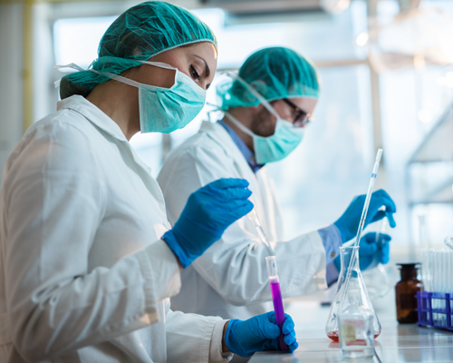 A photo of two researchers working in a lab. They are both wearing white lab coats, blue gloves, light blue hair nets and light blue face masks. The researcher in the foreground is holding a test tube in one hand and a pipette in the other. The person in the background is pouring liquid from a test tube in to a clear glass flask.