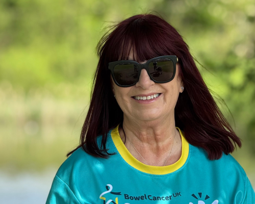 A photo of Julie smiling at the camera. She is wearing a light teal and yellow ActiveApril t-shirt and black sunglasses. Behind her is some greenery.