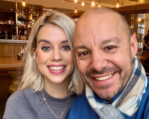 A photo of Charlie and her husband smiling at the camera. Charlie is on the left of the image and is wearing a grey top and a gold necklace. Her husband is on the left of the image and is wearing a black, tan and white plaid scarf and a blue top. In the background there is a bar.