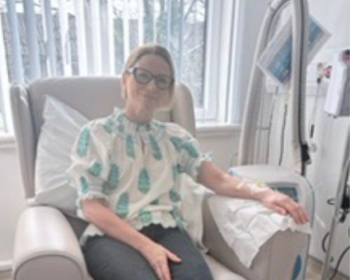 A photo of Mel sitting in a hospital treatment chair receiving medication. Medical tubing is connected to Mel's left arm and is attached to equipment beside the chair. Mel is wearing a white blouse with a ruffled collar and a light teal oval pattern, along with dark jeans. Her left hand arm rests on the chair armrest and the other on the lap.