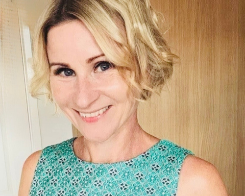 A selfie-style photo of Mel wearing a light teal sleeveless top with a dark blue four-leaf clover geometric pattern. Mel is smiling broadly and facing the camera, with short curly hair cropped around the cheekbones.