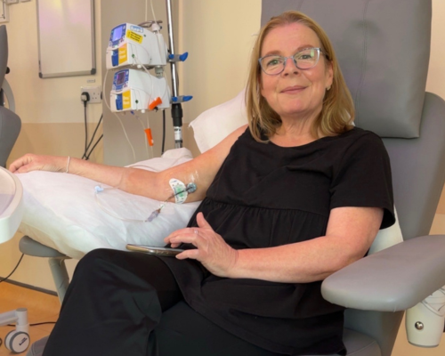 A photo of Jane sitting in a hospital treatment chair receiving medication. Her right arm rests on a cushion placed on top of the armrest, with medical tubing connected to her arm, extending to equipment behind her. Her phone is rested on her lap and she is using it with her left hand. Jane is wearing a loose black short sleeved top, and black trousers.