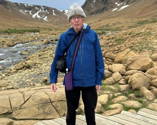 A photo of Jim standing on a wooden boardwalk with a snowy mountain landscape in the background. They're wearing a grey knitted beanie, rectangular black-framed glasses, black trousers and a dark blue coat zipped up. Two cross-body bags, one black and one purple, with a water bottle poking out of the purple bag.